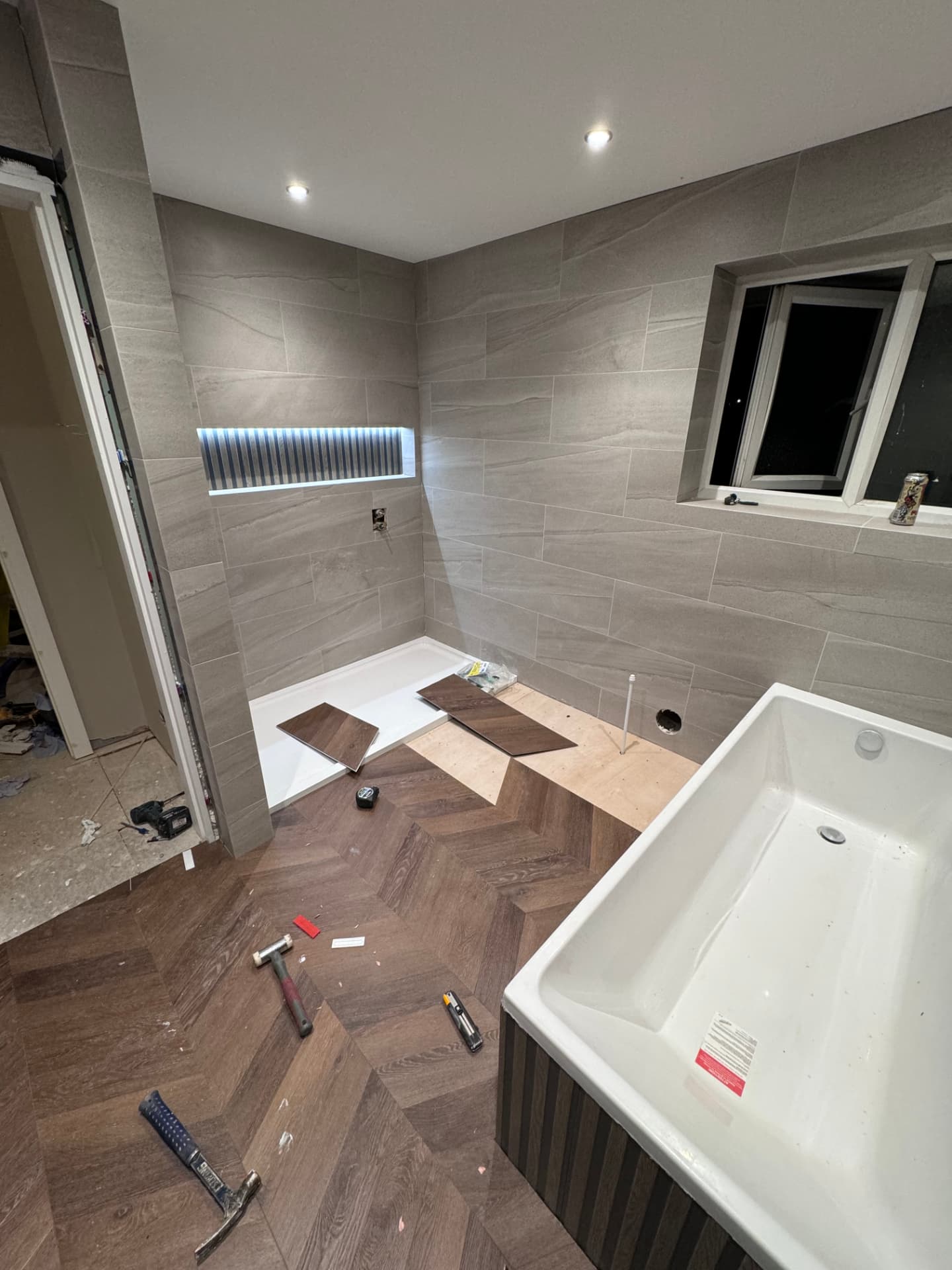 Family bathroom mid-fit with tiled walls, lit niche and herringbone flooring being laid